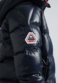 Marinblå puffjacka med en glänsande, vadderad yta och en hexagonal lapp med "JACKIT"-logotyp på vänstra ärmen. Dragkedjedetalj finns.