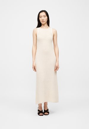 Femme debout, portant une robe en maille sans manches de couleur crème, longueur cheville, et des sandales à talons noires ouvertes sur un fond uni.