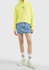 Neon gul hoodie med logotyp, denim minikjol med vågmönster, vita och neon gröna sneakers, i kombination med vita strumpor med varumärkesdesign.