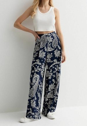 Žena v bílém crop topu a širokých námořnicky modrých kalhotách s bílým paisley vzorem, doplněná bílými teniskami, stojí před jednoduchým pozadím.