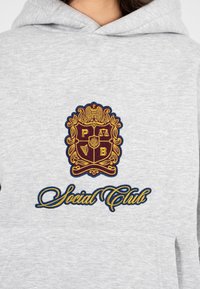 Felpa grigia con un grande emblema ricamato in rosso scuro e oro, con il testo "Social Club" in corsivo blu. Tasca a marsupio sotto.