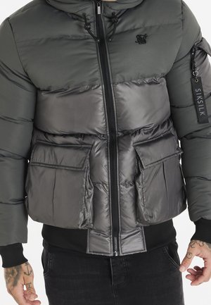 Veste matelassée gris foncé avec fermeture éclair frontale, grandes poches à rabat, poche zippée à la manche avec étiquette SikSilk, portée par une personne aux mains tatouées.