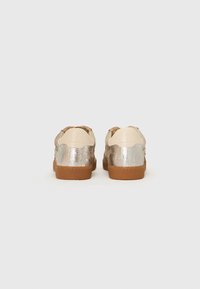 Sneakers with a beige, smooth leather upper and metallic silver accents. Textured rear panel and a brown rubber sole.