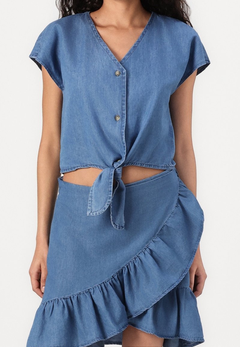 Femme portant un haut en denim bleu clair à manches courtes boutonné et noué à la taille, avec une jupe portefeuille à volants assortie, sur fond blanc.