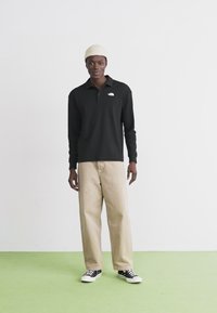 Svart langermet poloshirt med hvitt logo, beige vide bukser og svarte sneakers på en modell mot en ensfarget bakgrunn.