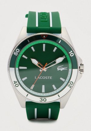 Green Lacoste wristwatch with silver markers, white and orange hands, green bezel with minute markings, and green strap with white stripes.