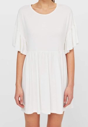 Frau trägt ein kurzes weißes Kleid mit rundem Ausschnitt, geraffter Taille und kurzen, flatternden Ärmeln vor einem einfarbigen Hintergrund.