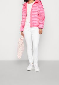 Różowa kurtka puchowa z kapturem, posiadająca diagonalne przeszycia i zamek błyskawiczny z przodu. W połączeniu z białymi dżinsami i wzorzystą torbą wielokrotnego użytku.
