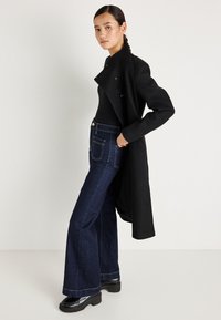 Fekete gyapjú kabát magas gallérral, sötétkék, harangszárú farmernadrággal, kontrasztvarrással és fekete lakkbőr cipővel párosítva.