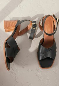 Schwarze Ledersandalen mit quadratischer Zehenpartie und über Kreuz verlaufenden Riemen. Mit einer hellbraunen Innensohle und einem strukturierten Blockabsatz aus Holz. Verstellbarer Knöchelriemen.