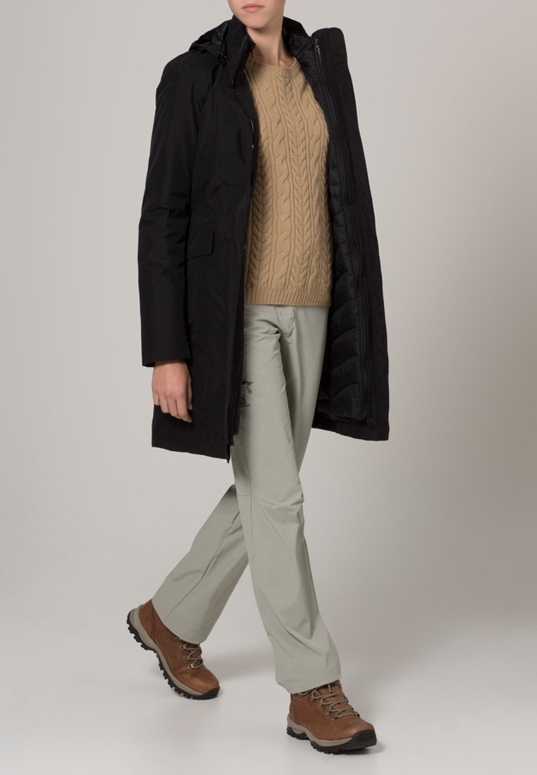 Parka noire mi-longue avec une fermeture éclair, de grandes poches et une capuche, portée sur un pull en maille torsadée beige et un pantalon clair avec des bottes de randonnée marron.