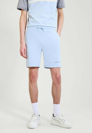 Mann trägt hellblaue Shorts mit Kordelzug, weiße Sportsocken und weiße Sneakers, steht mit einer Hand in der Tasche.
