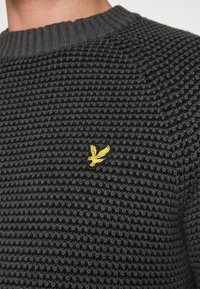 Knitted sweater in dark gray with a textured pattern. Features a ribbed neckline and a small yellow eagle logo on the chest.