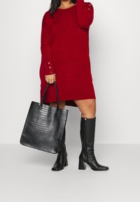 Robe-pull rouge côtelée avec des accents de boutons, associée à un sac fourre-tout noir texturé à motif crocodile et des bottes hautes noires à talons.