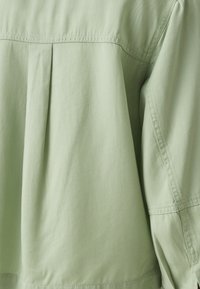 Primo piano di tessuto verde chiaro con cuciture visibili e una piega centrale sul pannello posteriore di un indumento.