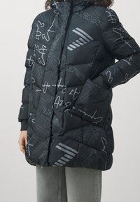 Kvinna i svart pufferjacka med abstrakta vita text- och logomönster, framdragkedja och sidofickor, kombinerad med grå jeans.