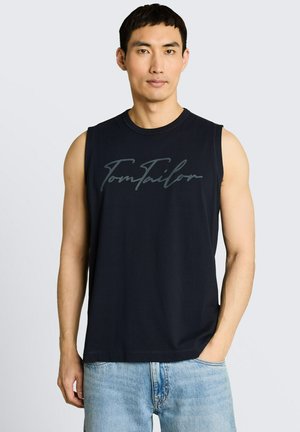 Man met een mouwloos zwart Tom Tailor-shirt en lichtblauwe spijkerbroek, staand met één hand in zijn zak tegen een effen achtergrond.