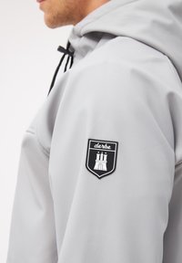 Hellgrauer Pullover-Hoodie mit schwarzen Akzenten. Verfügt über einen auffälligen schwarz-weißen Aufnäher am linken Ärmel. Glatte Stoffstruktur.