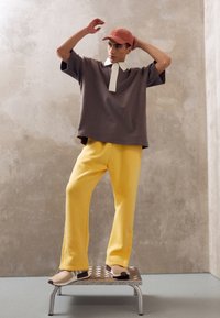 Baggy brun t-shirt med vit krage, ihop med klara gula byxor och beige sneakers. Modellen står på en silverplatta.