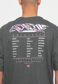 Retro della maglietta grigia scura con testo stilizzato "Originals European Tour" e un elenco di città europee in lettere bianche e rosa.