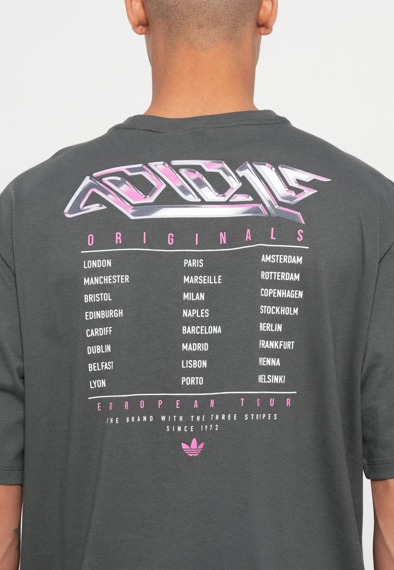 Parte posterior de una camiseta gris oscuro con el texto estilizado "Gira Europea Originals" y una lista de ciudades europeas en letras blancas y rosas.