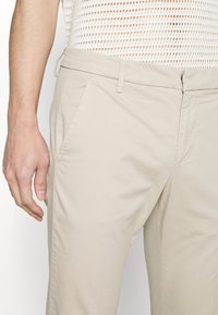 Pantalon en coton beige présentant une texture lisse, une coupe droite à l'avant, des poches latérales, des passants pour ceinture, et une coupe ajustée, montré de la taille au genou.
