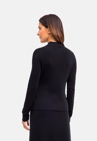 Frau mit glattem braunem Haar, die einen engen schwarzen Pullover und einen schwarzen Rock trägt, von hinten vor einem schlichten hellen Hintergrund.