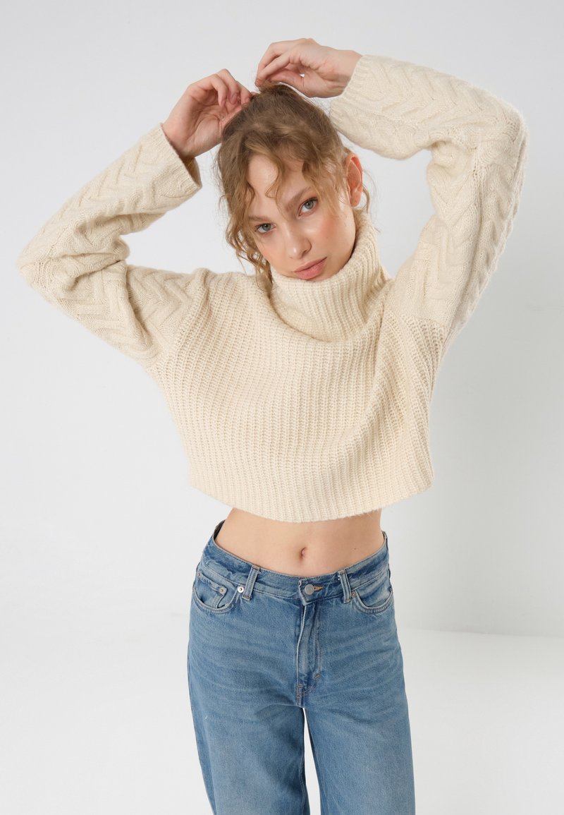 Cropped, cream-colored knit sweater with a high turtleneck, featuring a textured pattern, paired with high-waisted blue jeans.