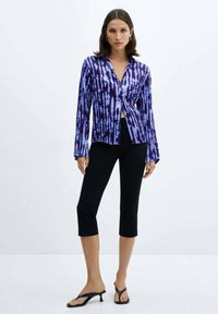 Vrouw in een lila-paarse en blauwe tie-dye blouse met lange mouwen, zwarte cropped broek en zwarte hakken, staand tegen een plain witte achtergrond.