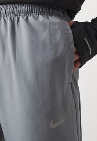 Gros plan d'une personne portant un pantalon gris de Nike avec une taille élastique et la main dans la poche latérale, habillée d'un haut noir à manches longues avec une bande réfléchissante.