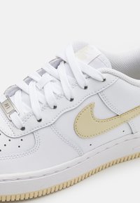 Fehér sneaker sima bőr felsőrésszel, arany Nike logóval, fehér fűzős, perforált orrrésszel és textúrált gumi talppal.