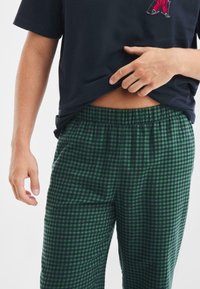 Pantalones de pijama a cuadros verdes y azul marino con cintura elástica, combinados con una camiseta negra que presenta un pequeño diseño bordado.