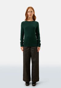 Dunkelgrüner Langarmpullover mit Rundhalsausschnitt, kombiniert mit hochgeschnittenen, weit geschnittenen dunkelblauen Jeans und schwarzem Schuhwerk.