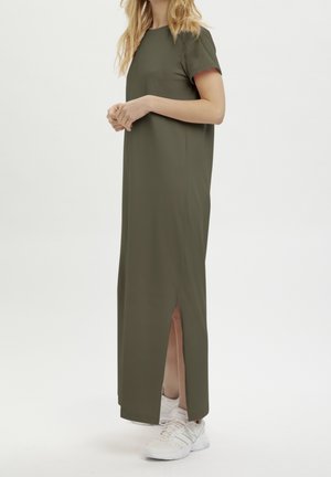 Femme portant une robe longue à manches courtes de couleur vert olive avec une fente latérale et des baskets blanches, debout devant un fond clair uni.