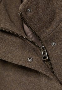 Gros plan d'une veste en laine marron montrant une fermeture éclair ouverte et des boutons-pression métalliques au niveau du col.