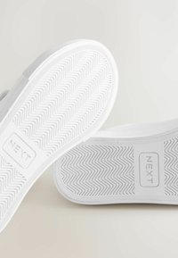 Vita gummisneakersolar med zigzag-mönster och en rektangulär etikett präglad med "NEXT." Texturerat material med en slät yta.