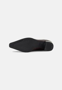 Chaussure habillée à bout pointu avec semelle noire, couchée sur le côté, montrant une semelle en caoutchouc texturée et un talon empilé sur fond blanc.