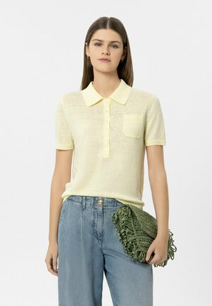 Frau mit glattem braunem Haar, die ein hellgelbes kurzärmeliges Hemd mit Kragen und hoch taillierte blaue Jeans trägt, hält eine grüne geflochtene Clutch.