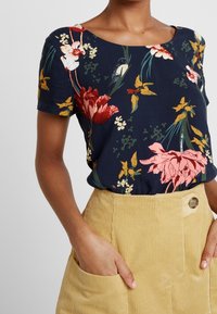 Donna che indossa una camicia floreale blu navy a maniche corte infilata in pantaloni di velluto a coste beige a vita alta con bottone e tasche.