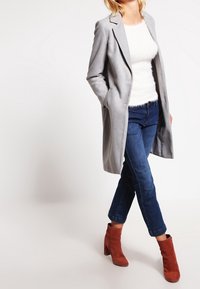 Abrigo largo gris claro con solapas muestradas, combinado con una blusa blanca esponjosa, jeans azules y botas de tobillo color óxido. Texturas suaves en todo.