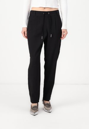Black tapered trousers with a drawstring waist, pleats at the front, and side pockets. Paired with metallic silver sneakers.