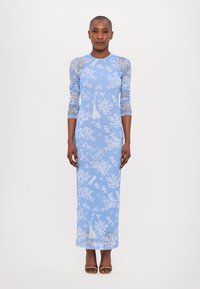 Robe longue à manches longues de couleur bleue avec des motifs floraux, tissu semi-transparent, coupe ajustée et détails subtils en pampilles. Associée à des talons bruns.