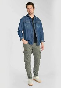 Jeanshemd mit geknöpften Brusttaschen, darüber getragen ein schwarzes T-Shirt; grüne Cargo-Hose mit Reißverschluss-Details; beige Sneaker.