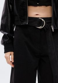 Μαύρο δερμάτινο κοντό μπουφάν με γυαλιστερή υφή, συνδυασμένο με  μαύρο ψηλόμεσο παντελόνι που διαθέτει ασημένια αγκράφα ζώνης σε σχήμα κύκλου και πιέτες.