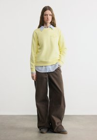 Νέα γυναίκα που στέκεται φορώντας κίτρινο φούτερ πάνω από μπλε ριγέ πουκάμισο, φαρδιά καφέ παντελόνια και γκρι αθλητικά παπούτσια σε γκρι δάπεδο.