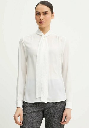 Donna che indossa una blusa bianca a maniche lunghe con una sciarpa annodata al collo e pantaloni grigi con trama, in piedi davanti a uno sfondo chiaro e uniforme.