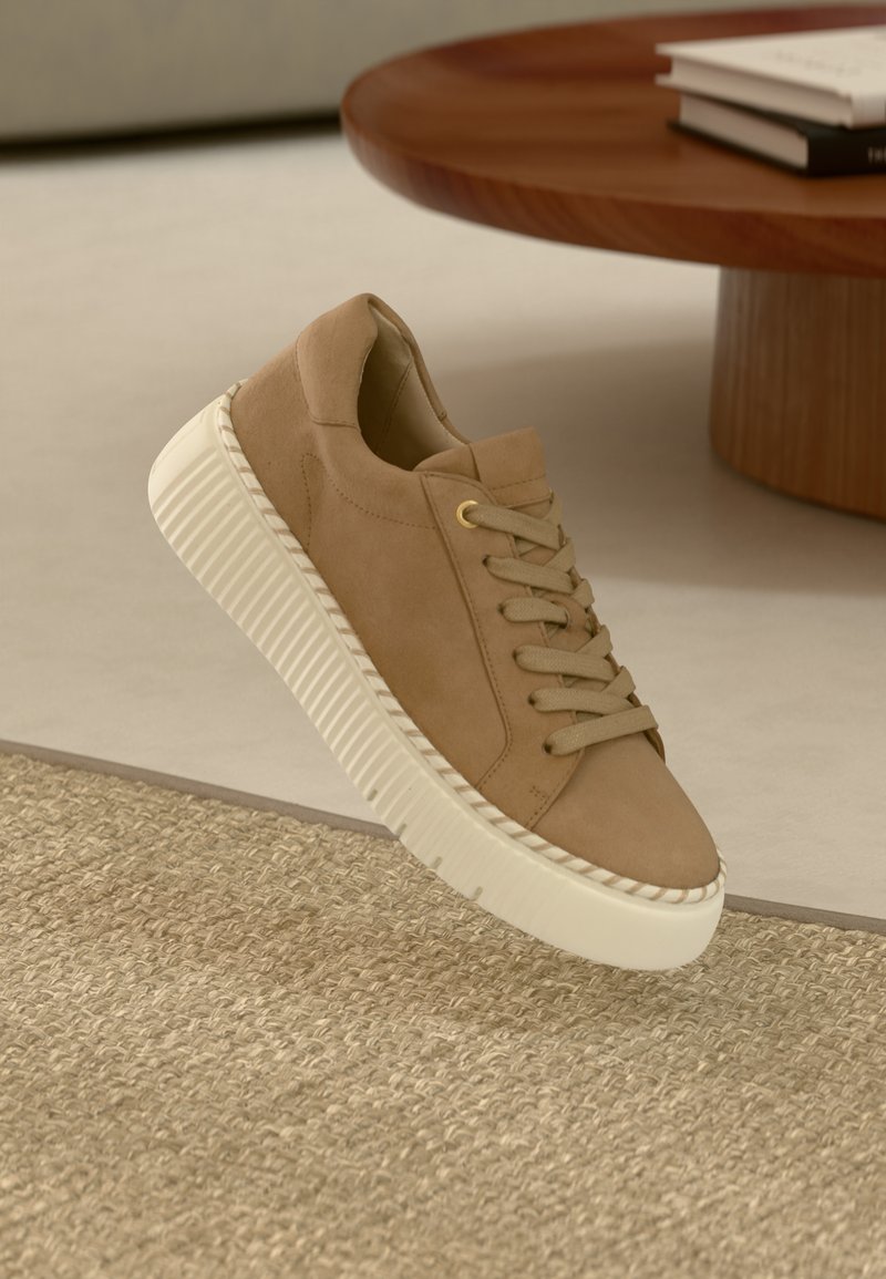 Béžový semišový teniska s texturovanou, mimo-bílou gumovou podrážkou, s kulatými tkaničkami a prošitým designem podél okraje podrážky.