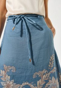 Blauwe linnen rok met bloemende borduursel in lichtbeige en bruin. Heeft een strik taille met gouden hardware accenten aan de uiteinden.