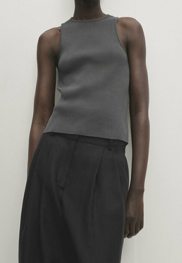Personne portant un haut gris sans manches côtelé et un pantalon noir ample à taille haute, debout sur un fond clair uni.