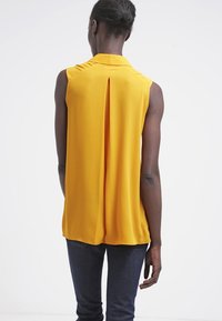 Bezramenná žlutá blůza s límcem a skladem vzadu, vyrobená z hladké, lehké textilie. Nosená s tmavými džínami.